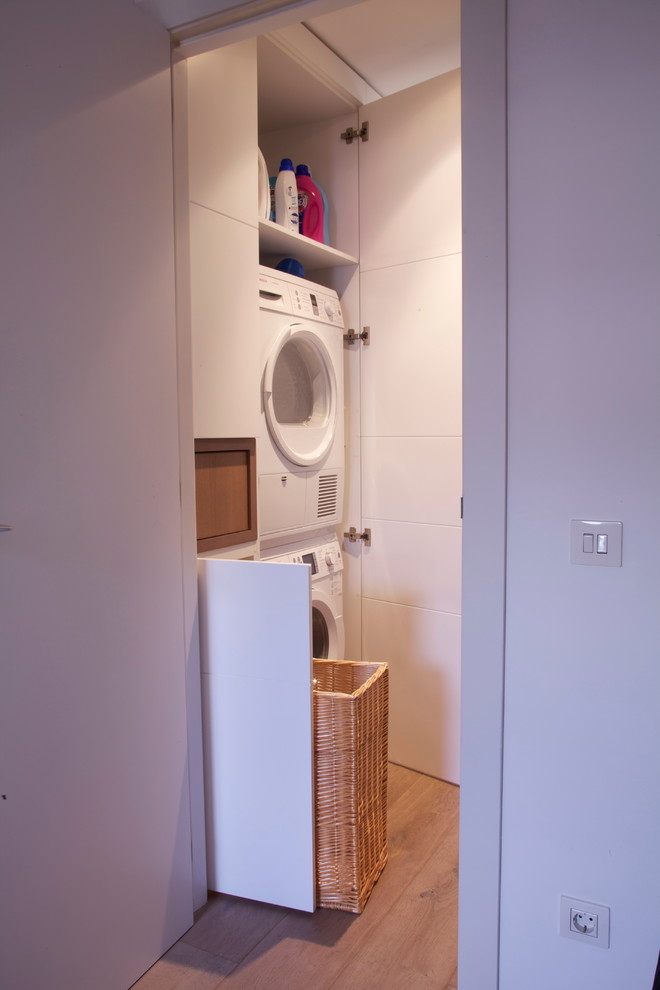 Kleiner Moderner Hauswirtschaftsraum mit Waschmaschinenschrank, flächenbündigen Schrankfronten, weißen Schränken, weißer Wandfarbe, braunem Holzboden und Waschmaschine und Trockner gestapelt in Barcelona