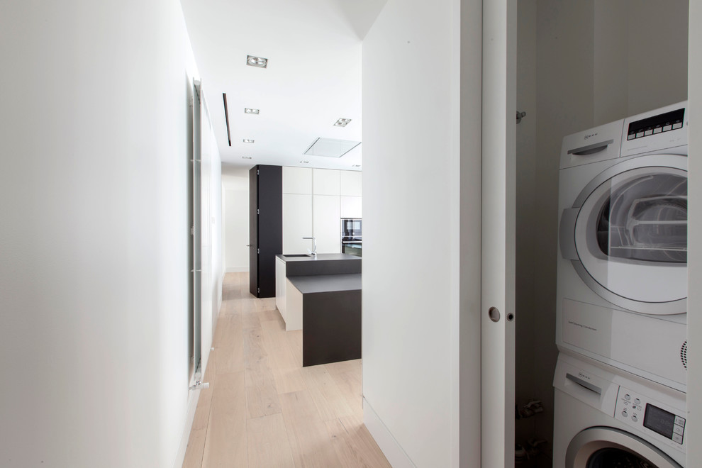 Inspiration for a small modern single-wall laundry cupboard in Madrid with flat-panel cabinets, white cabinets, white walls, light hardwood flooring and a stacked washer and dryer.