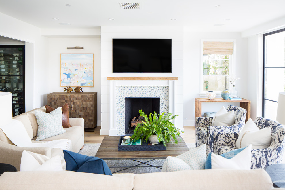 На фото: гостиная комната в морском стиле с белыми стенами, светлым паркетным полом, стандартным камином, фасадом камина из плитки, телевизором на стене и бежевым полом
