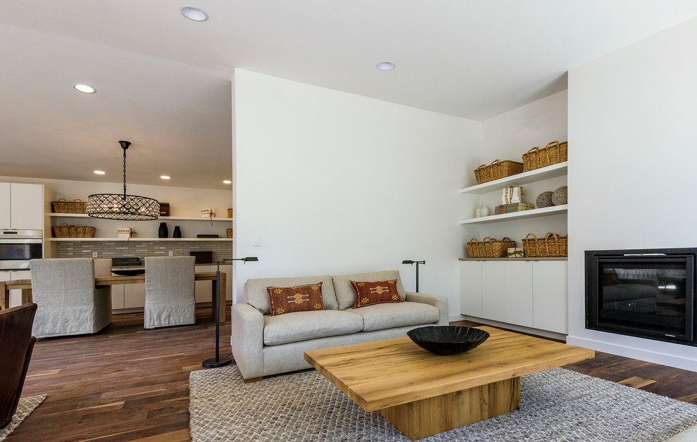 Cette image montre un petit salon minimaliste ouvert avec un mur blanc, un sol en bois brun, une cheminée standard, un manteau de cheminée en plâtre, un téléviseur fixé au mur et un sol marron.