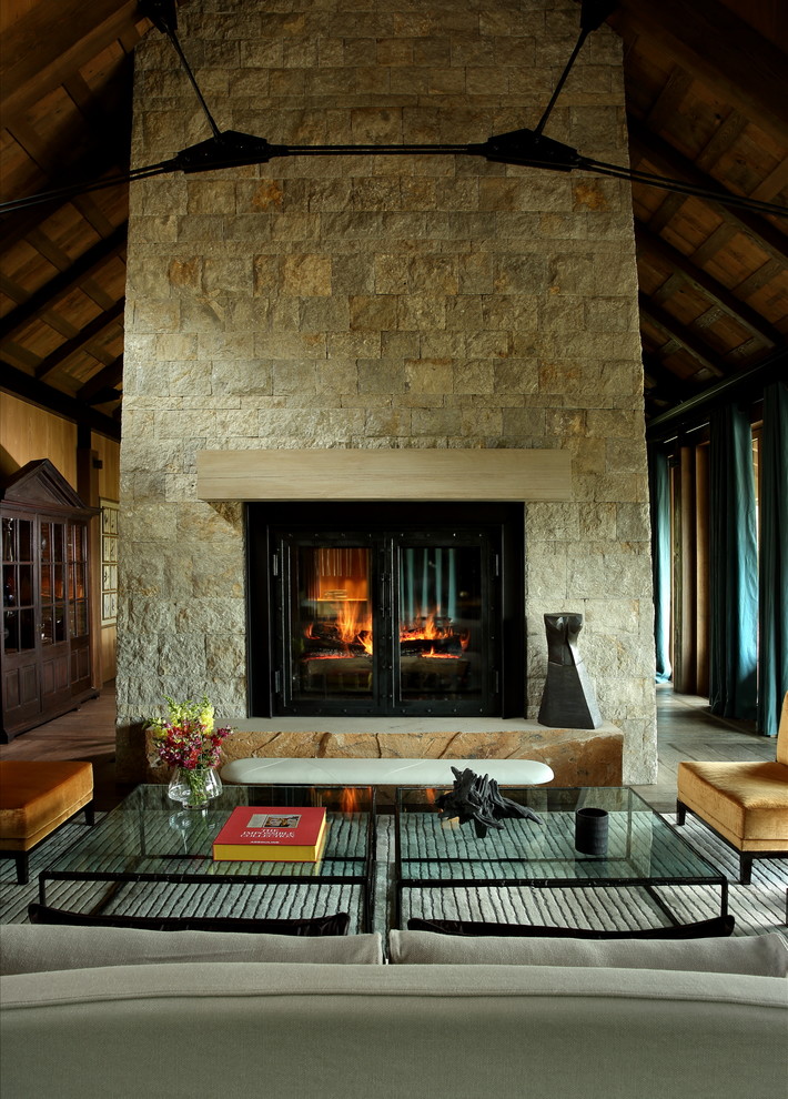 Ejemplo de salón abierto rural grande con paredes marrones, suelo de madera en tonos medios, chimenea de doble cara, marco de chimenea de piedra y suelo gris