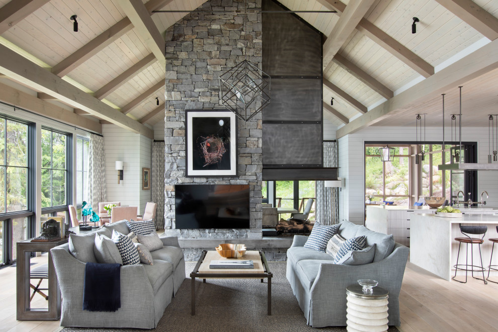 Imagen de salón abierto rural con chimenea de doble cara, televisor colgado en la pared, paredes blancas, suelo de madera clara, marco de chimenea de piedra y suelo beige