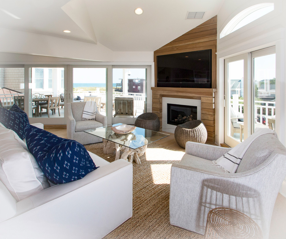 Imagen de salón abierto costero grande con paredes blancas, suelo de madera clara, chimenea de esquina, marco de chimenea de madera y televisor colgado en la pared