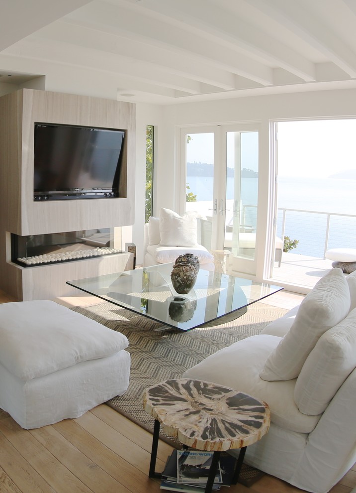 Ejemplo de salón para visitas abierto costero grande con paredes blancas, suelo de madera clara, chimenea lineal, marco de chimenea de madera y televisor retractable