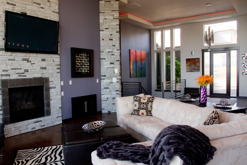 На фото: огромная открытая гостиная комната в современном стиле с фиолетовыми стенами, темным паркетным полом, стандартным камином, фасадом камина из камня и телевизором на стене