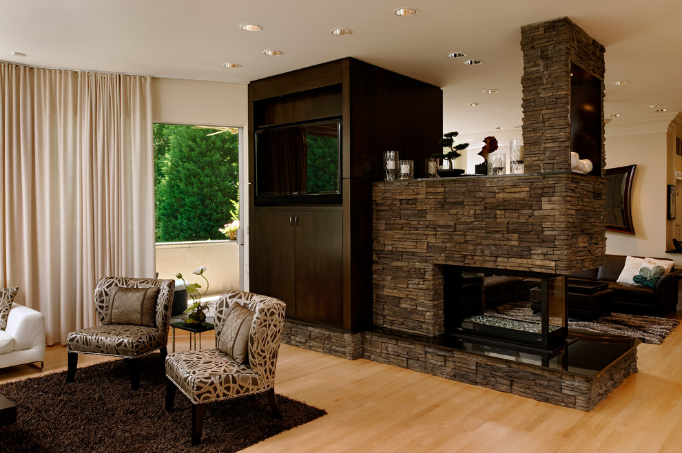 Modelo de salón contemporáneo con paredes beige, suelo de madera clara, chimenea de doble cara, marco de chimenea de piedra y pared multimedia