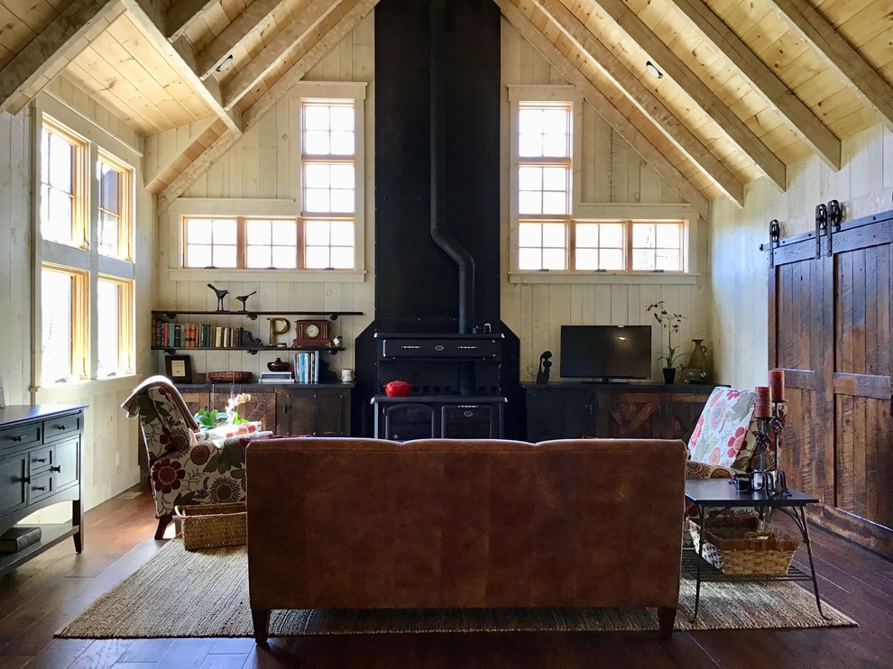 Modelo de salón abierto de estilo de casa de campo de tamaño medio con paredes beige, suelo de madera oscura, estufa de leña, televisor independiente y suelo marrón
