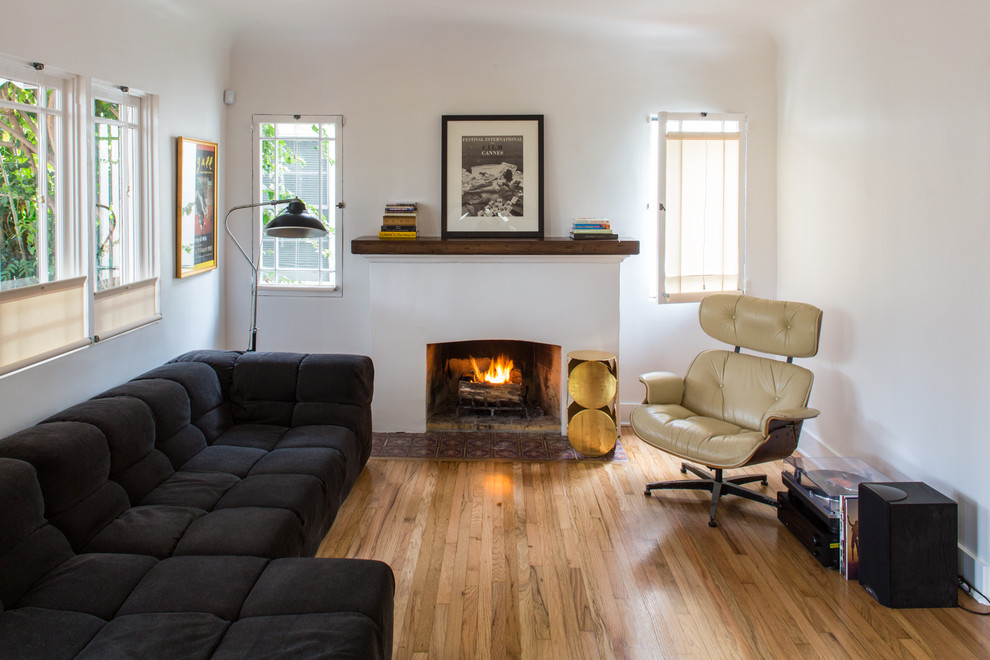 ロサンゼルスにある中くらいなコンテンポラリースタイルのおしゃれなリビング (標準型暖炉、白い壁、淡色無垢フローリング、漆喰の暖炉まわり、黒いソファ) の写真
