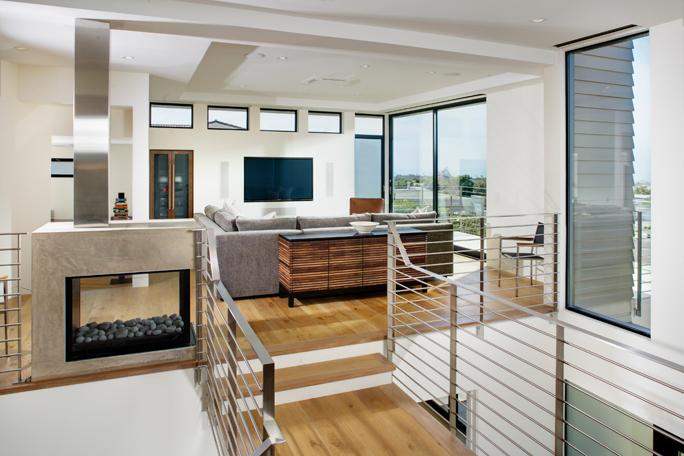 Modelo de salón abierto contemporáneo de tamaño medio con paredes blancas, suelo de madera clara, chimenea de doble cara, televisor colgado en la pared y suelo beige