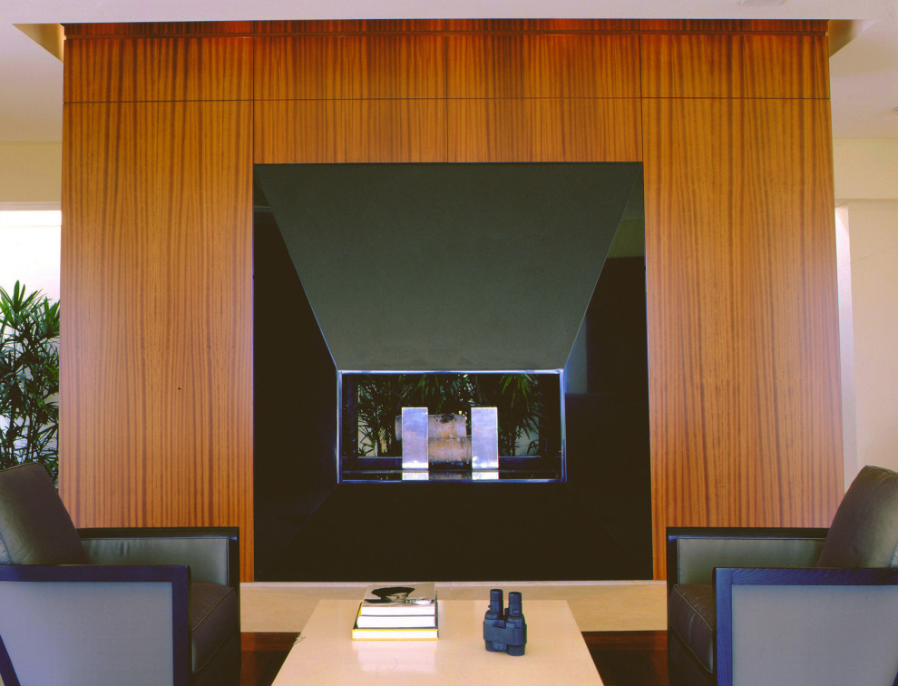 На фото: большая открытая гостиная комната в современном стиле с с книжными шкафами и полками, коричневыми стенами, полом из известняка, стандартным камином, фасадом камина из дерева и бежевым полом