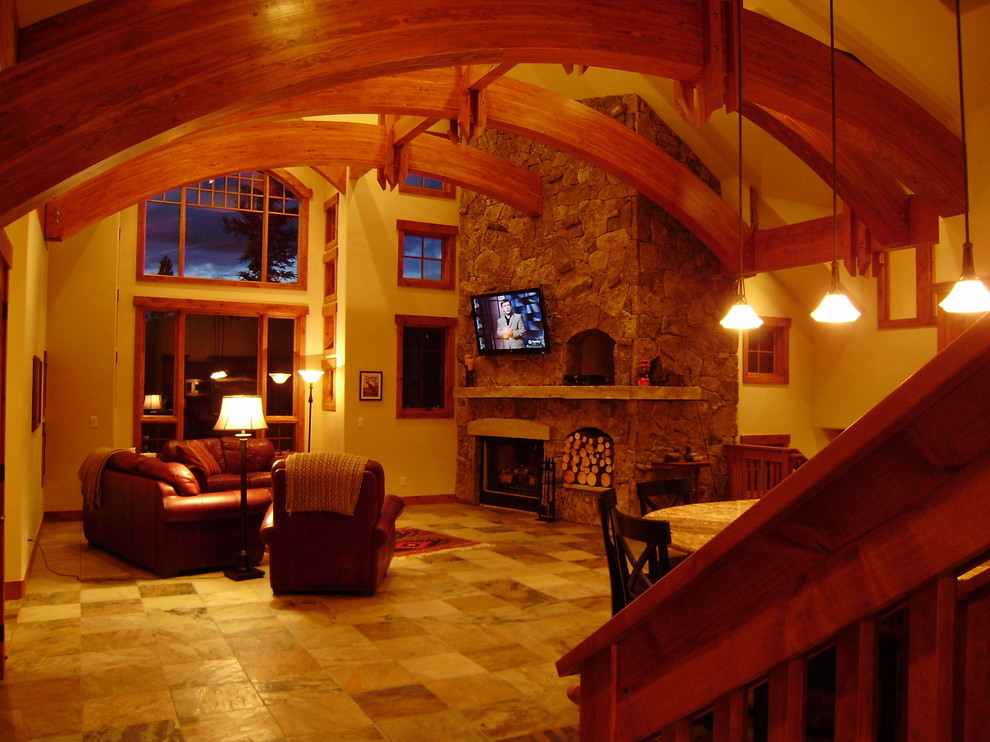 Cette photo montre un grand salon montagne ouvert avec un sol en ardoise, une cheminée standard, un manteau de cheminée en pierre, un téléviseur fixé au mur, un sol beige et un mur beige.