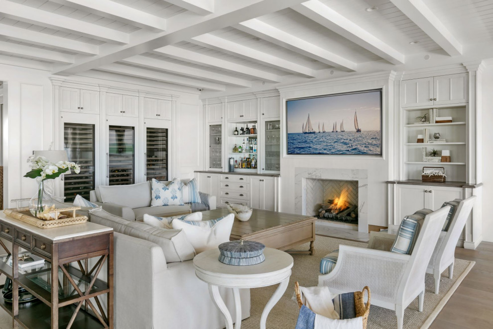 Cette image montre un grand salon marin ouvert avec un bar de salon, un mur blanc, un sol en bois brun, une cheminée standard, un manteau de cheminée en pierre, un téléviseur fixé au mur et un sol gris.