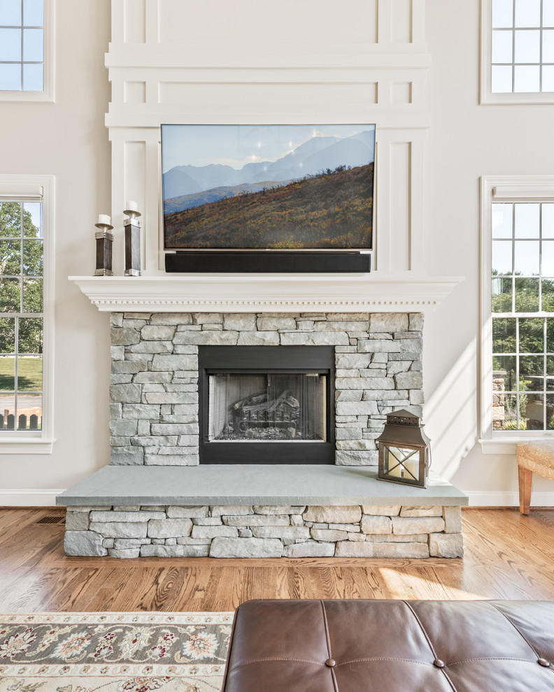 This is an example of a large classic enclosed living room in Philadelphia with white walls, medium hardwood flooring, a standard fireplace, a stone fireplace surround, a wall mounted tv and brown floors.