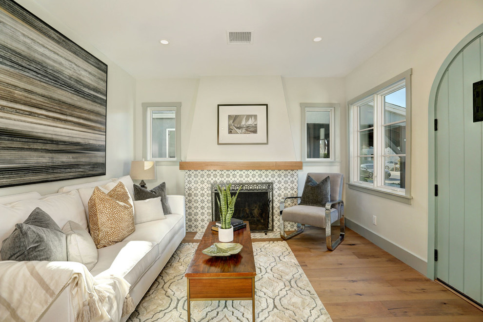 На фото: гостиная комната в стиле неоклассика (современная классика) с белыми стенами, паркетным полом среднего тона, стандартным камином, фасадом камина из плитки и коричневым полом без телевизора с