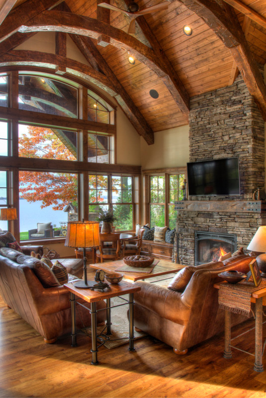 Large mountain style enclosed medium tone wood floor and brown floor living room photo in Minneapolis with beige walls, a standard fireplace, a stone fireplace and a wall-mounted tv