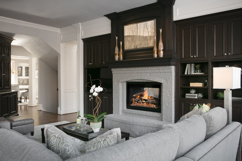 Diseño de salón abierto clásico renovado grande con paredes grises, suelo de madera en tonos medios, chimenea de doble cara, marco de chimenea de ladrillo y pared multimedia