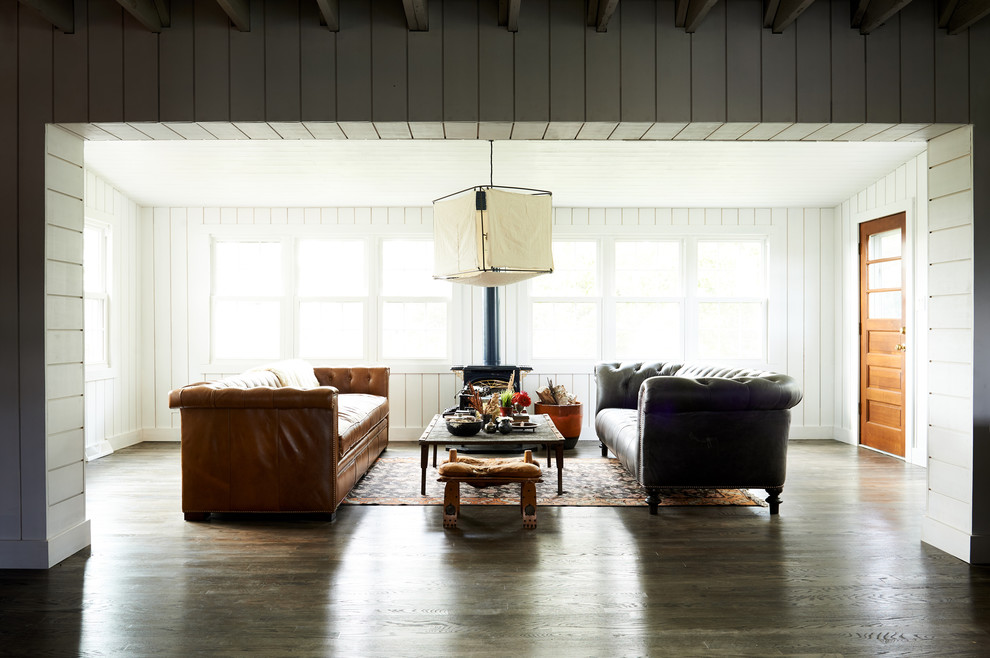 Modelo de salón abierto moderno sin televisor con paredes blancas, suelo de madera oscura, estufa de leña, marco de chimenea de metal y suelo marrón