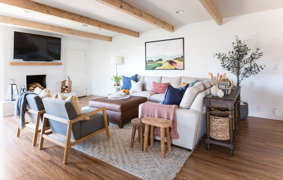 Inspiration for a medium sized farmhouse open plan living room in Phoenix with white walls, laminate floors, a standard fireplace, a brick fireplace surround, a wall mounted tv, brown floors and exposed beams.