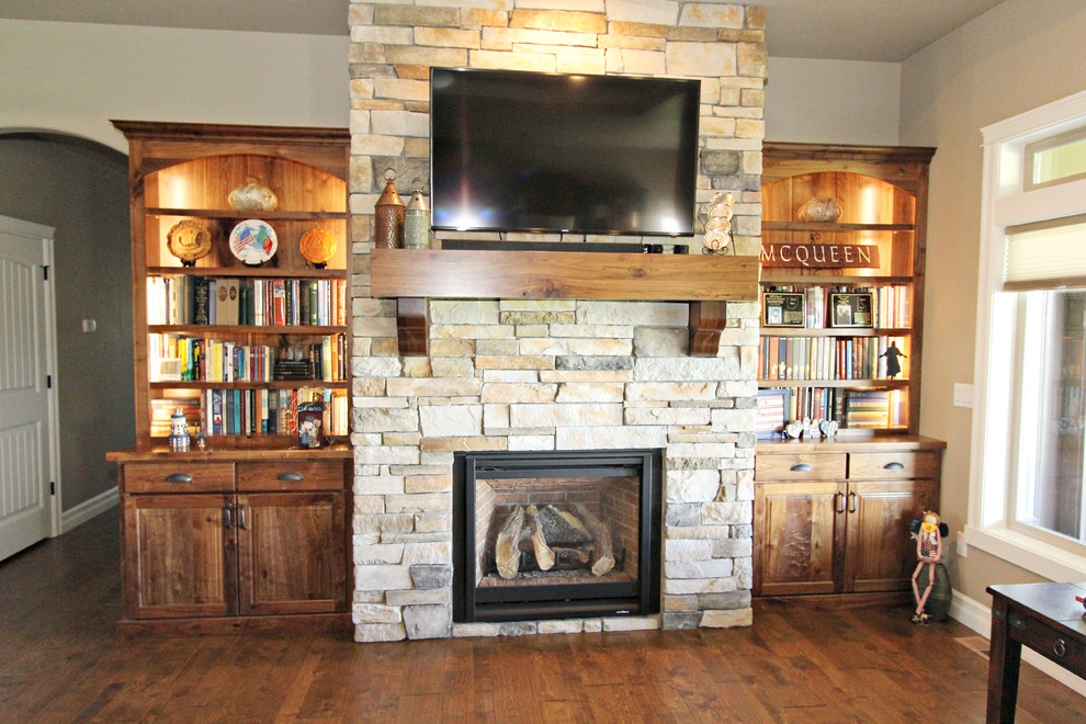 Cette photo montre un grand salon craftsman ouvert avec un mur beige, un sol en bois brun, une cheminée standard, un manteau de cheminée en pierre et un téléviseur fixé au mur.