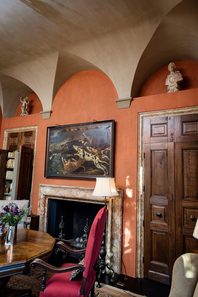 Cette image montre un grand salon méditerranéen fermé avec une bibliothèque ou un coin lecture, un mur orange, parquet foncé, une cheminée standard et un manteau de cheminée en pierre.