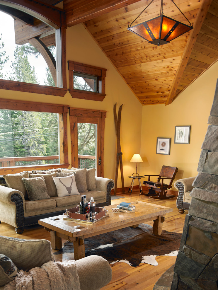 Imagen de salón abierto rural grande con paredes amarillas, suelo de madera en tonos medios y marco de chimenea de piedra
