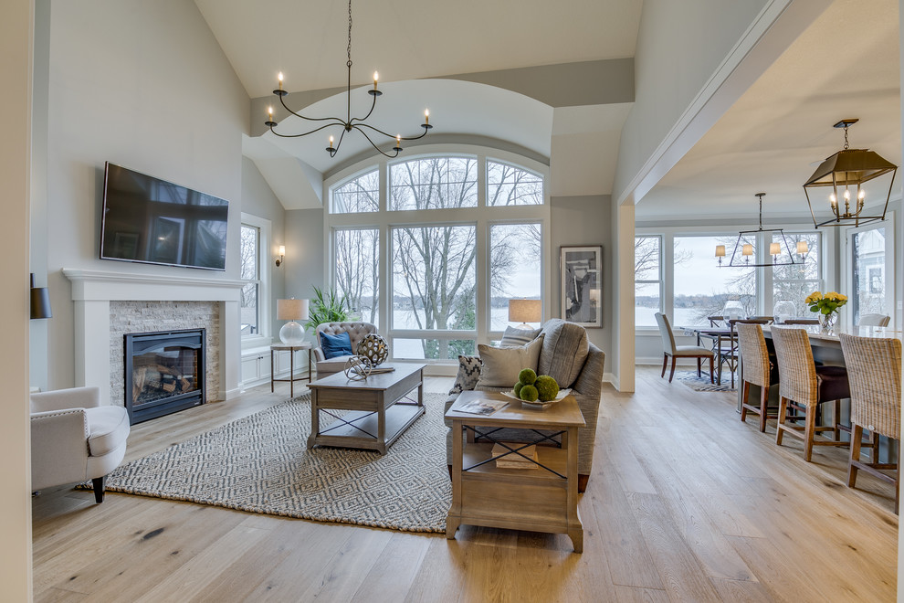 This is an example of a large rural open plan living room in Minneapolis with grey walls, light hardwood flooring, a standard fireplace, a brick fireplace surround, a wall mounted tv and beige floors.