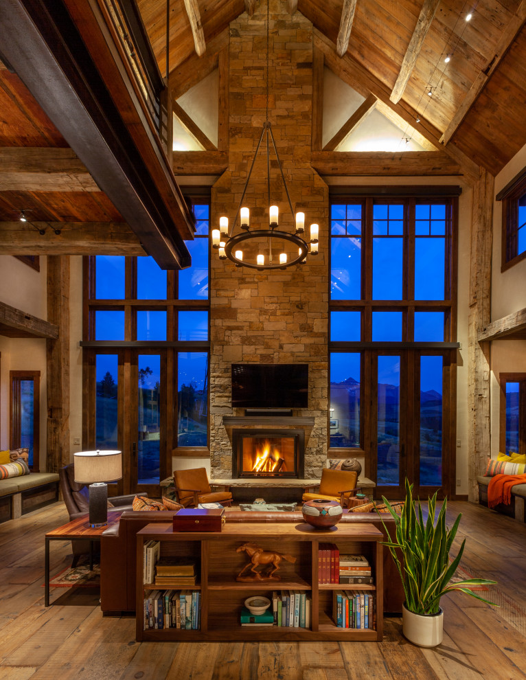 This is an example of a rustic open plan living room in Denver with beige walls, medium hardwood flooring, a standard fireplace, a stacked stone fireplace surround, a wall mounted tv, brown floors, exposed beams, a vaulted ceiling and a wood ceiling.