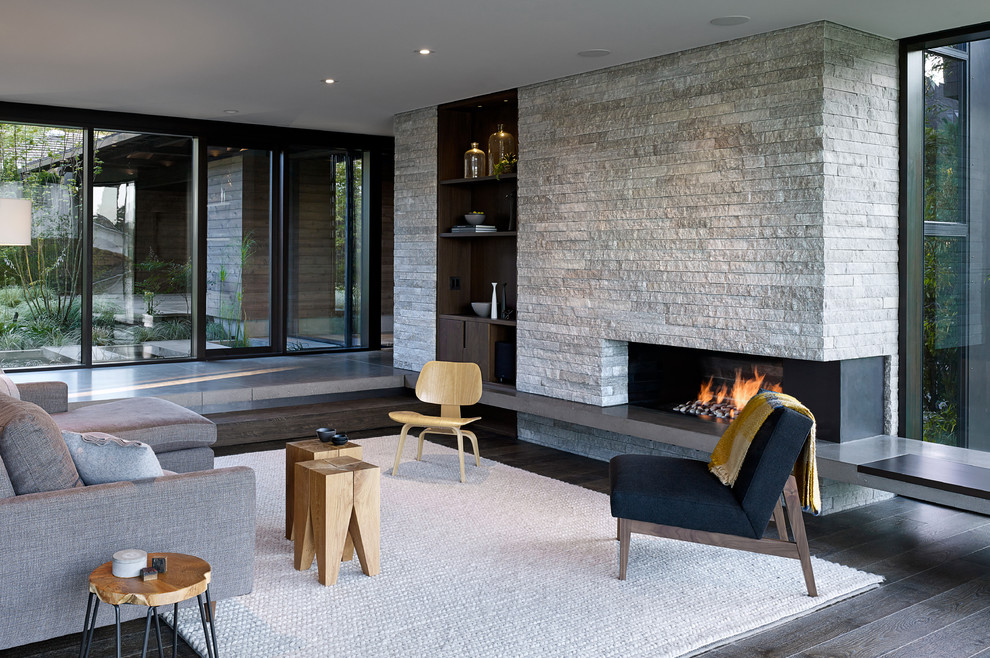 Imagen de salón abierto actual con suelo de madera oscura, chimenea lineal, marco de chimenea de piedra, suelo marrón y alfombra