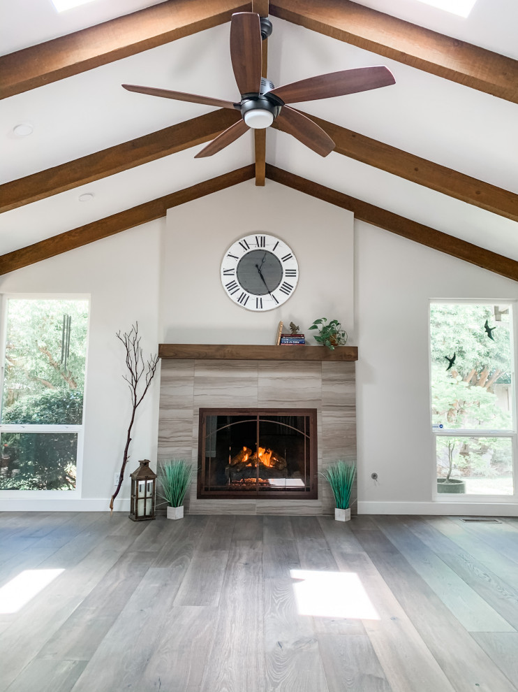 Cette photo montre un grand salon nature ouvert avec un mur gris, un sol en bois brun, une cheminée standard, un manteau de cheminée en carrelage, un sol gris et poutres apparentes.