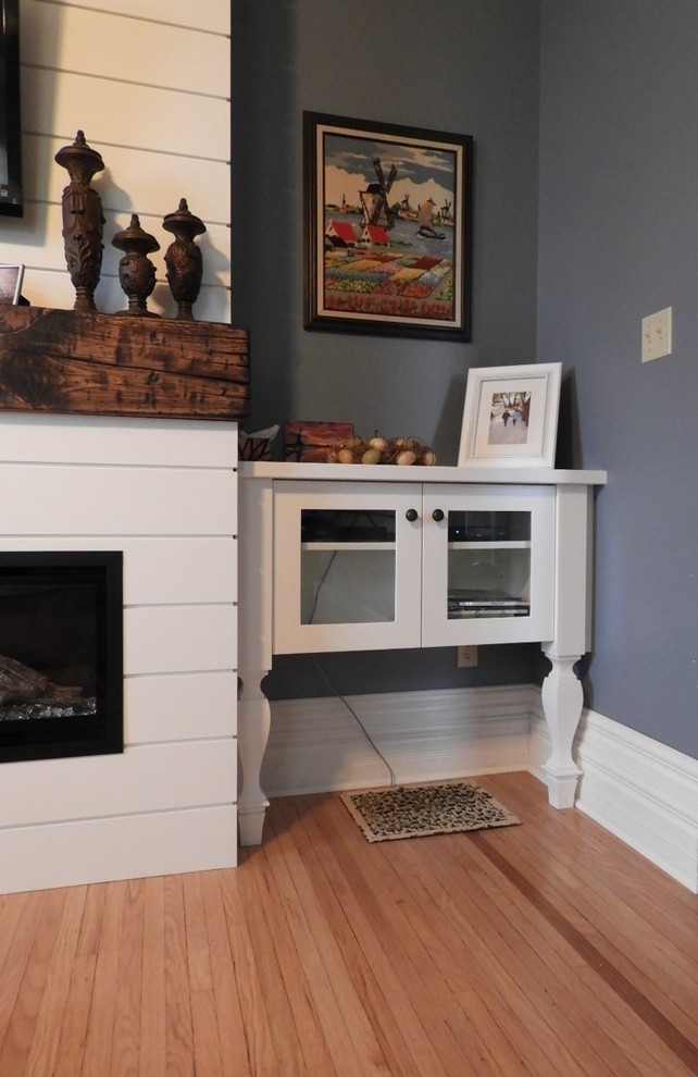 This is an example of a small farmhouse enclosed living room in Toronto with blue walls, light hardwood flooring, a hanging fireplace, a wooden fireplace surround, a wall mounted tv and brown floors.