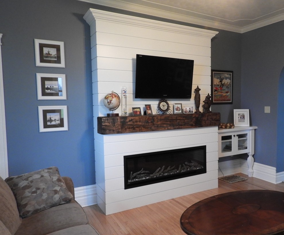 トロントにあるお手頃価格の小さなカントリー風のおしゃれな独立型リビング (青い壁、淡色無垢フローリング、吊り下げ式暖炉、木材の暖炉まわり、壁掛け型テレビ、茶色い床) の写真