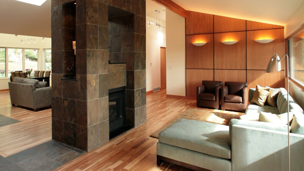 Imagen de salón abierto actual grande con paredes beige, suelo de madera clara, chimenea de doble cara y marco de chimenea de piedra