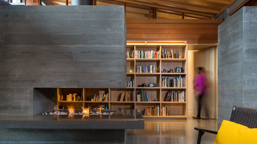 Esempio di un soggiorno minimalista stile loft con libreria, pareti grigie, camino bifacciale, cornice del camino in cemento, nessuna TV e soffitto in legno