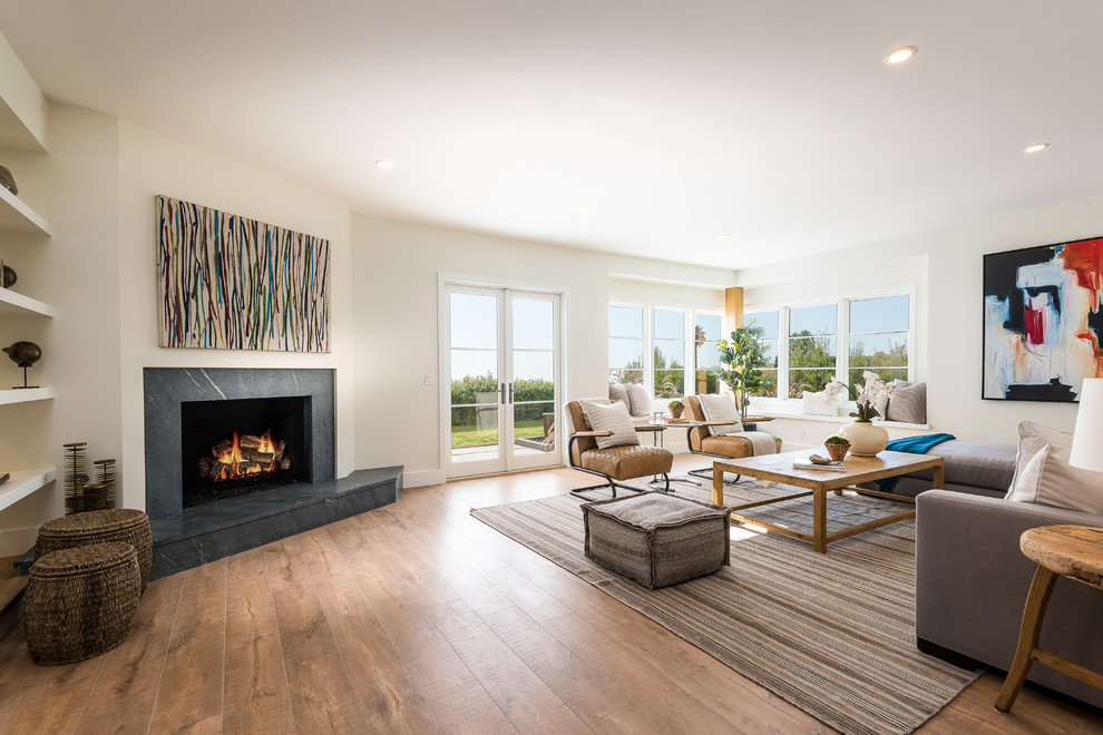 This is an example of a beach style living room in Los Angeles with white walls, medium hardwood flooring, a corner fireplace and a stone fireplace surround.