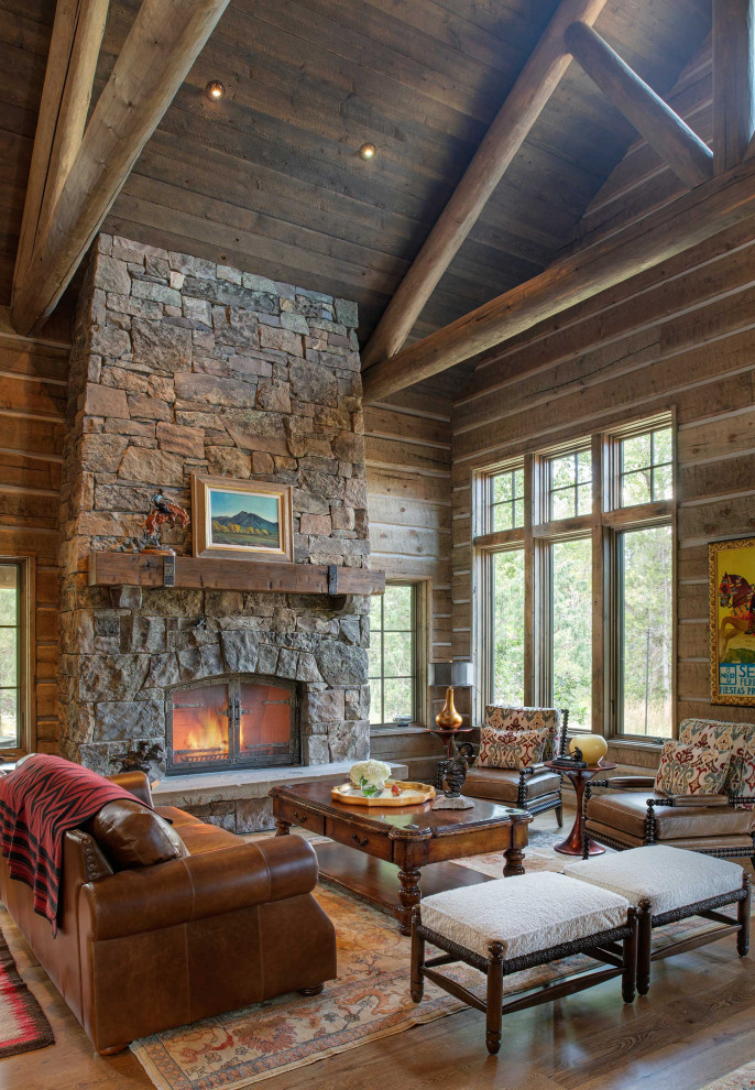 Exemple d'un salon montagne en bois ouvert avec un mur marron, un sol en bois brun, une cheminée standard, un manteau de cheminée en pierre, un sol marron, poutres apparentes, un plafond voûté et un plafond en bois.