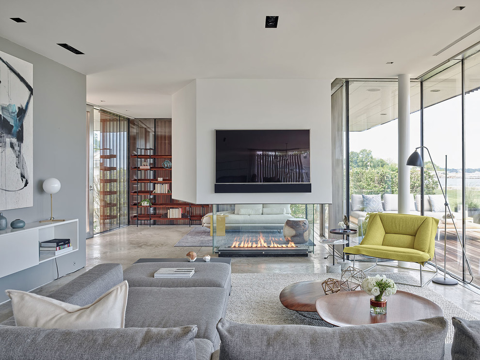 Imagen de salón abierto actual de tamaño medio con paredes blancas, suelo de cemento, chimenea de doble cara, televisor colgado en la pared y alfombra