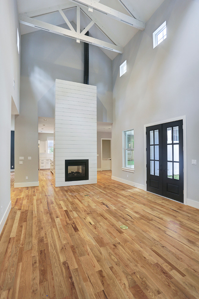 Cette image montre un grand salon rustique ouvert avec un mur gris, parquet clair, une cheminée double-face, un manteau de cheminée en bois, une salle de réception, aucun téléviseur et un sol marron.