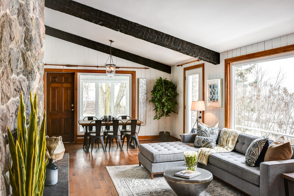 Medium sized farmhouse open plan living room with white walls, medium hardwood flooring, a standard fireplace, a stone fireplace surround, no tv, brown floors, exposed beams, a vaulted ceiling and panelled walls.