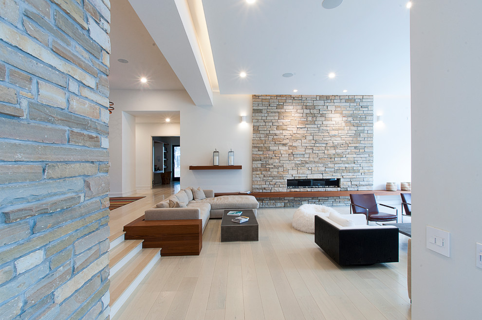 Modelo de salón abierto contemporáneo grande sin televisor con paredes blancas, suelo de madera clara y marco de chimenea de piedra