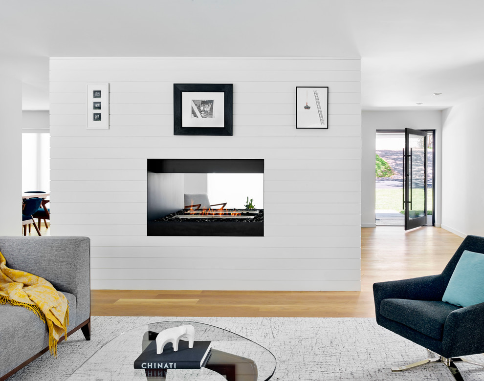 Cette photo montre un salon rétro ouvert avec un mur blanc, parquet clair, une cheminée double-face, un manteau de cheminée en bois et un téléviseur dissimulé.