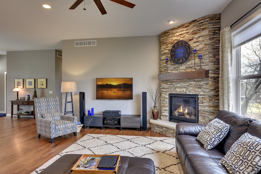 Foto de salón de estilo americano con paredes grises, suelo de madera en tonos medios, chimenea de esquina, marco de chimenea de piedra y televisor colgado en la pared