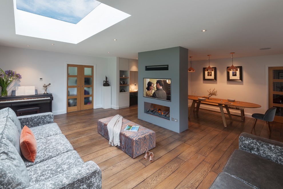 Example of a large trendy open concept medium tone wood floor and brown floor living room design in Surrey with a bar, gray walls, a two-sided fireplace, a plaster fireplace and a wall-mounted tv