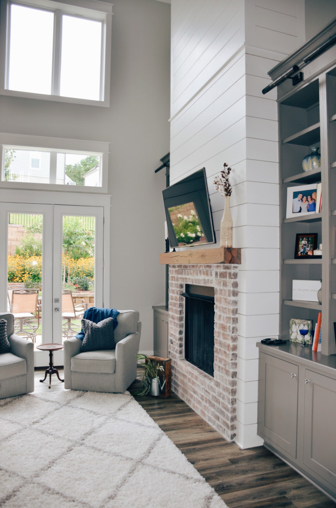 На фото: открытая гостиная комната в стиле кантри с серыми стенами, светлым паркетным полом, стандартным камином, фасадом камина из кирпича, телевизором на стене и серым полом с