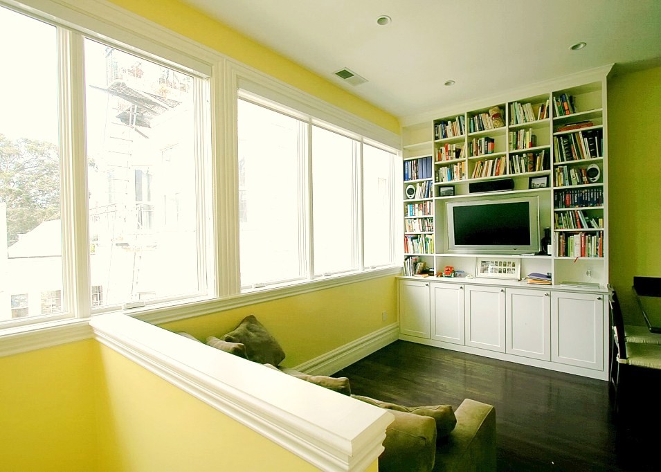 Modern inredning av ett stort separat vardagsrum, med gula väggar, mörkt trägolv, en väggmonterad TV och ett bibliotek