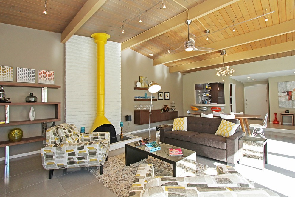 Cette photo montre un grand salon gris et jaune rétro ouvert avec un mur beige, un sol en carrelage de céramique, un poêle à bois, un manteau de cheminée en brique, un sol gris et un téléviseur fixé au mur.