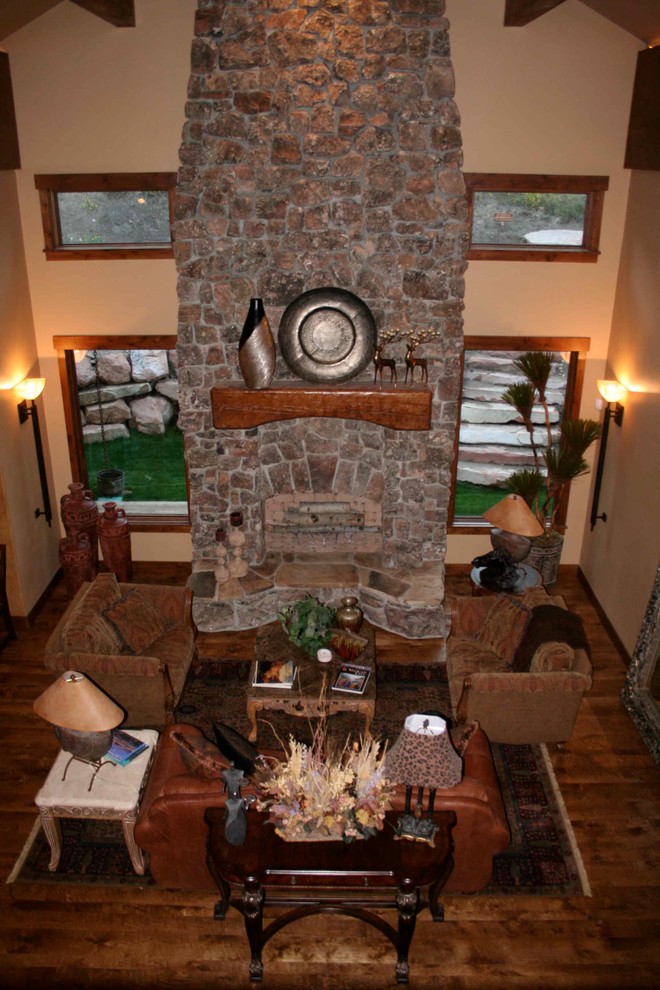 Cette photo montre un grand salon montagne ouvert avec une salle de réception, un mur beige, un sol en bois brun, une cheminée standard et un manteau de cheminée en pierre.