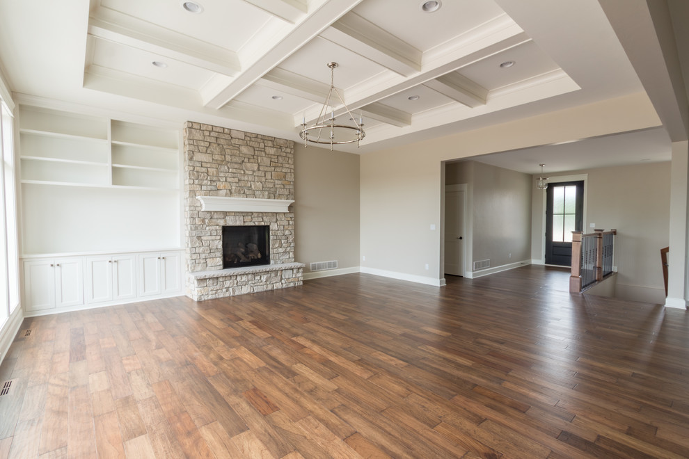 Cette image montre un grand salon rustique ouvert avec un mur gris, un sol en bois brun, une cheminée standard, un manteau de cheminée en pierre, un téléviseur encastré et un sol marron.