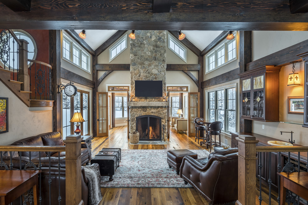 Cette photo montre un grand salon montagne ouvert avec un bar de salon, un mur beige, un sol en bois brun, une cheminée double-face, un manteau de cheminée en pierre et un sol marron.