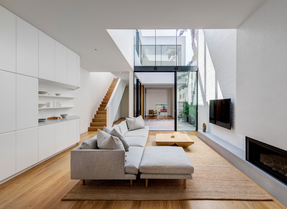 Стильный дизайн: гостиная комната в современном стиле с белыми стенами, паркетным полом среднего тона, горизонтальным камином, телевизором на стене и коричневым полом - последний тренд