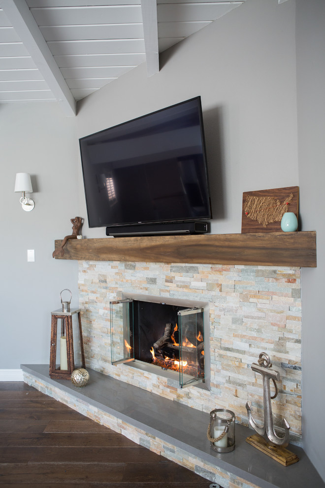 Modelo de salón abierto costero de tamaño medio con paredes grises, suelo de madera en tonos medios, chimenea de esquina, marco de chimenea de piedra y televisor colgado en la pared